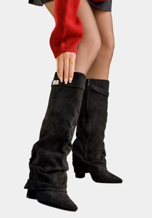 Jambes en bottes hautes en suède noir avec une boucle, portant des collants transparents et un pull rouge, main ajustant le haut de la botte gauche.