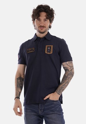 Uomo con capelli ricci che indossa una polo blu navy con toppe ricamate e jeans, in piedi con una mano in tasca.