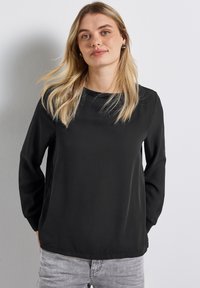 Černý top s dlouhým rukávem z hladkého materiálu, s kulatým výstřihem a volným střihem. Obsahuje jemné švy a mírně zakřivený lem.