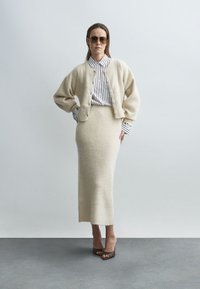 Cardigã de lã cor creme com mangas largas, com botões à frente, usado por cima de uma camisa às riscas e de uma saia comprida combinando. Sandálias castanhas de salto.