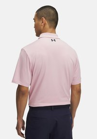 Hellrosa Polohemd mit Kragen und kurzen Ärmeln, versehen mit einem kleinen schwarzen Logo auf der Rückseite und strukturiertem Stoff. Getragen mit einer marineblauen Hose.