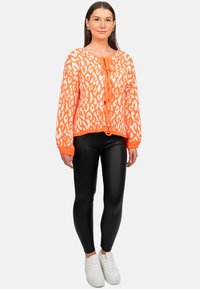 Maglione lavorato a maglia arancione brillante con macchie leopardate bianche, dotato di chiusura a cravatta sul davanti e polsini a costine, abbinato a leggings neri e sneaker bianche.