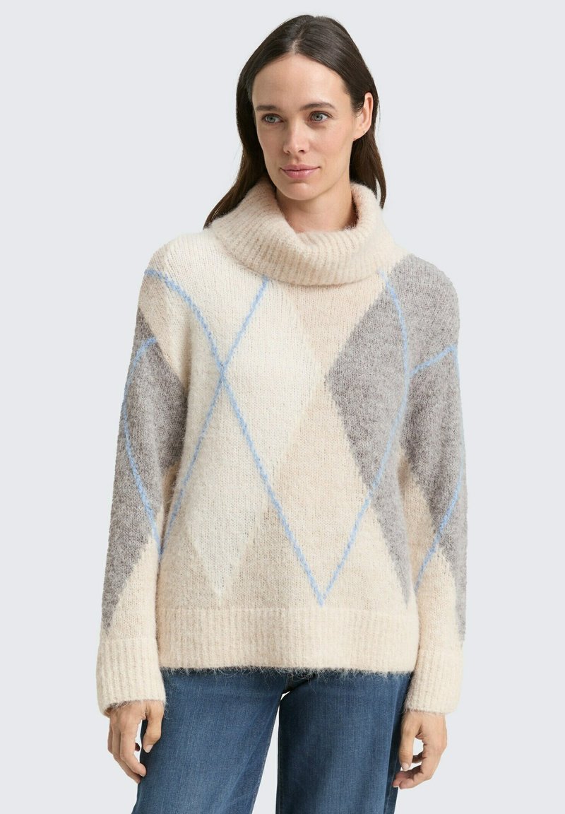 Femme portant un pull col roulé à motif argyle crème et gris avec des lignes bleues, associé à un jean bleu, se tenant devant un fond uni.