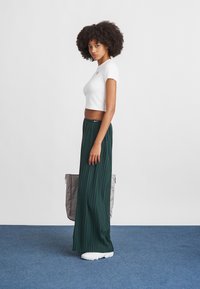 Top cropped branco, calças largas pregueadas verdes e sapatos brancos. O modelo está de lado contra um fundo plano, segurando uma bolsa cinza.