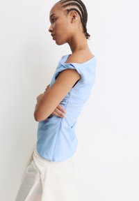 Unge person med flettet hår iført en lys blå off-shoulder top og cremefarvede bukser, stående sidelæns med armene korslagte mod en hvid baggrund.