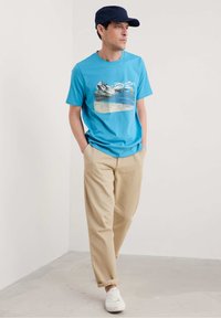 Mėlyna medvilninė marškinė su grafiniu paplūdimio atvaizdu, derinami su šviesiai rudos spalvos laisvo kirpimo kelnėmis ir baltomis sportbačiais.