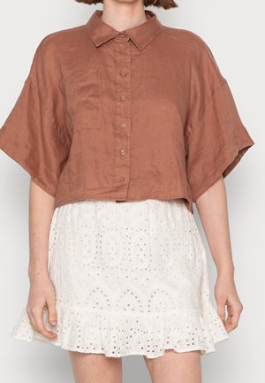 Femme portant une chemise marron ample à boutons avec des manches courtes et une jupe blanche en dentelle à œillets avec un ourlet volanté.