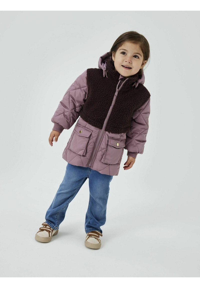 Padded Jacke in Mauve mit einem dunkelbraunen, flauschigen oberen Bereich, ausgestattet mit einem Reißverschluss vorne, Kapuze und zwei vorderen Taschen. Getragen mit blauen Jeans und Turnschuhen.