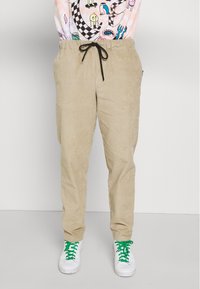 Béžové kordové kalhoty s elastickým pasem, s texturovanou tkaninou, bočními kapsami a volným střihem, v kombinaci s bílými teniskami a zelenými tkaničkami.