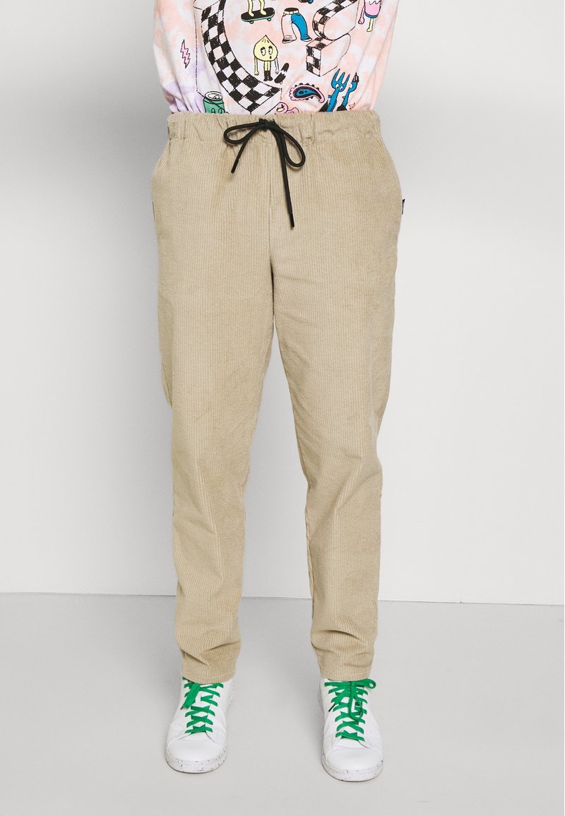 Béžové kordové kalhoty s elastickým pasem, s texturovanou tkaninou, bočními kapsami a volným střihem, v kombinaci s bílými teniskami a zelenými tkaničkami.
