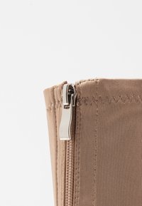 Primer plano de una tela beige con un zipper de metal plateado parcialmente cerrado sobre un fondo blanco.