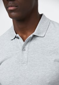 Jasnoszara koszulka polo wykonana z fakturowanej tkaniny z dwoma guzikami przy kołnierzyku. Zbliżenie na dekolt i obszar ramienia.