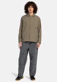 Khaki lightweight jacket with a collared design, front pocket, and subtle stitching details, paired with gray baggy pants and black shoes.