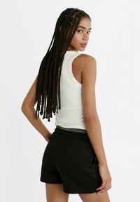 Kvinde med langt flet hår iført en hvid ærmeløs top og sorte shorts, som står med ryggen lidt væk mod en ensfarvet baggrund.