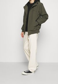 Olivgrüner Zip-Up-Jacke mit Kapuze, elastischen Bündchen und Seitentaschen. Trägt eine schwarze Bluse über creamfarbigen, lässig geschnittenen Hosen und schwarzen Turnschuhen.