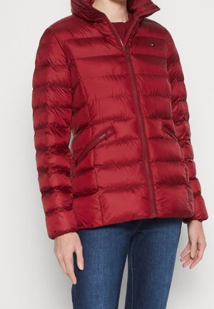 Vrouw die een rode doorgestikte pufferjas draagt met ritszakken en een blauwe spijkerbroek, staand tegen een effen achtergrond.