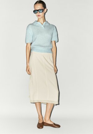 Femme portant un haut en maille bleu à manches courtes, une jupe midi beige, des ballerines marron et des lunettes de soleil foncées, debout devant un fond clair uni.