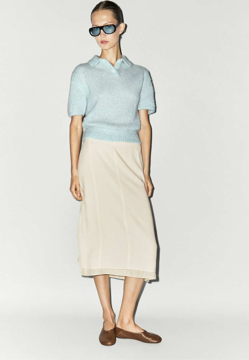 Woman wearing blue short-sleeve knit top, beige midi skirt, brown flats, and dark sunglasses standing against a plain light background.