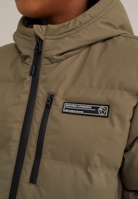 Oliivinvihreä puhvijakku, jossa on vetoketjukiinnitys ja korkea kaulus. Mukana musta etiketti valkoisella tekstillä sekä koripallografiikka.