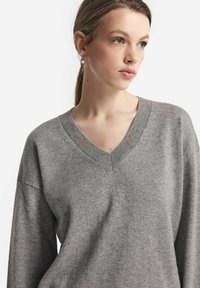 Szary sweter z dekoltem w serek i długimi rękawami, o miękkiej fakturze i prążkowanym wykończeniu wokół dekoltu, charakteryzujący się luźnym krojem i minimalistycznym stylem.