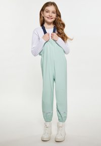 Jauna mergaitė su ilgais plaukais, vilkinti šviesiai žalius vandeniui nepralaidžius kombinezonus, baltą ilgomis rankovėmis marškinį ir baltus šnuromis batus prieš paprastą foną.