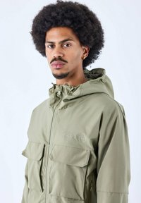 Giacca khaki con cappuccio in tessuto testurizzato, due ampie tasche frontali e dettaglio del logo. Presenta un cappuccio regolabile con cordino e chiusura a zip.