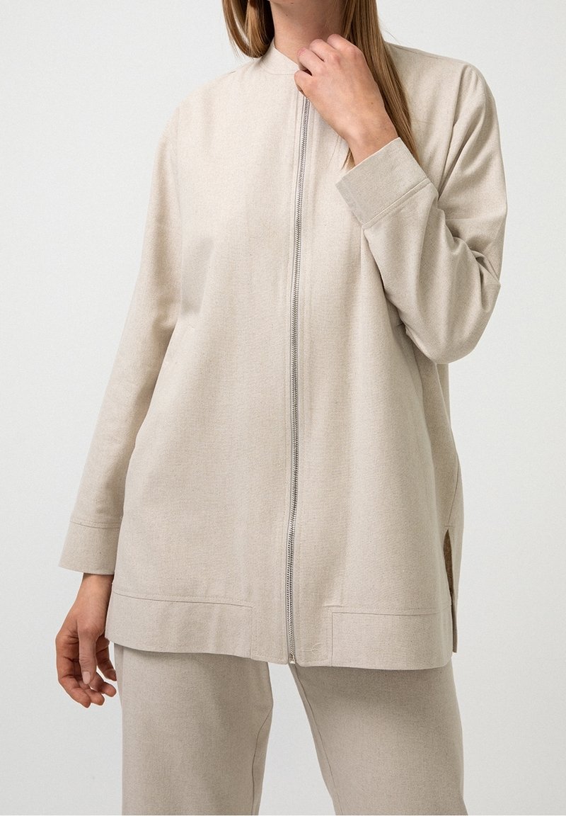 Bežová zipová bunda vyrobená z měkké tkaniny, s vysokým límcem, dlouhými rukávy a bočními rozparky na spodním okraji pro snadnější pohyb.