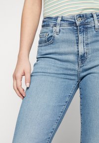 Gros plan sur une personne portant un jean en denim bleu clair et une chemise à rayures, avec une main reposant près de la cuisse.