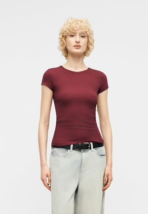 Ξανθός άτομο με σγουρά μαλλιά, φορώντας εφαρμοστό βυσσινί κοντομάνικο πουκάμισο και ανοιχτό μπλε τζιν με μαύρη ζώνη, στέκεται μπροστά σε απλό φόντο.