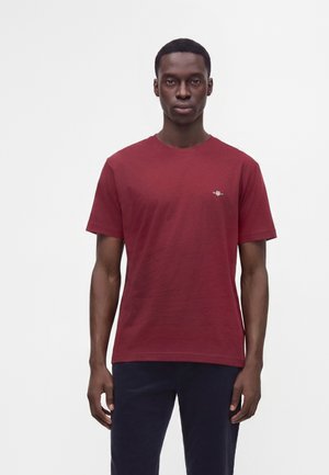 Mann mit kurzen Haaren, der ein schlichtes rotes Kurzarm-T-Shirt und dunkle Hosen trägt, vor einem schlichten weißen Hintergrund stehend.
