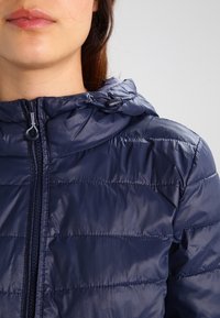 Námořnická modrá péřová bunda s kapucí, která obsahuje prošívané segmenty a přední zip, vyrobená z lesklého, lehkého materiálu.