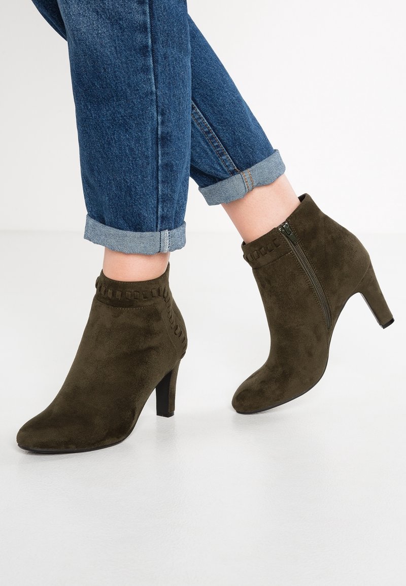 Bottes en daim vert olive montantes à la cheville avec détails de couture et fermeture éclair latérale, portées avec un jean bleu retroussé sur un sol blanc.