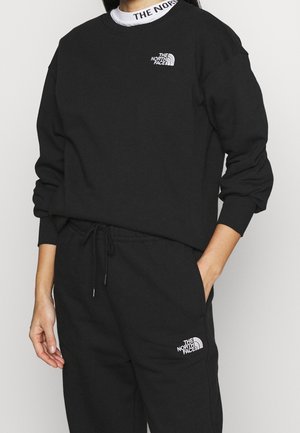 Personne portant un sweat-shirt noir The North Face et un pantalon de survêtement, avec les logos visibles sur la poitrine, le pantalon et le col d'un sous-pull blanc.