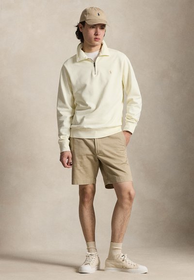 Jeune homme portant une casquette beige, un sweat-shirt crème à fermeture éclair au quart, un short kaki, des chaussettes beige et des baskets blanc cassé, debout avec une main dans la poche.