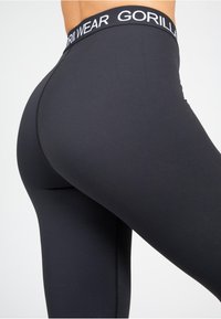 Crne tajice s širokim pojasom na kojem stoji "GORILLA WEAR" u bijelom tekstu. Tkanina izgleda glatko i elastično, s prilagođenim siluetom.