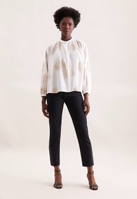 Weiße Bluse mit beigefarbenen Blumenmustern und gerafftem Ausschnitt, kombiniert mit schwarzen Skinny-Jeans und schwarzen, riemchenverzierten High Heels. Glatte Stofftextur.