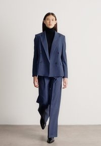 Sieviete pastaigājas tumsnē zila dubultpogas uzvalkā, melnā augstajā apģērbā un melnās smailās kurpēs uz balta fona.