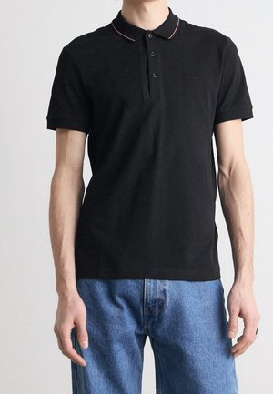 Sort poloshirt med en kontrasterende rød krave, forsynet med en fire-knaps knappelukning og et diskret logo på brystet. Bæres med lyseblå jeans.