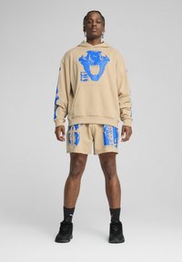 Beige hættetrøje med blå grafisk print, kengurulomme; matchende beige shorts med blå mønstre; båret med sorte sneakers, som viser atletisk fit.