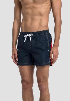 Torace maschile che indossa pantaloncini da bagno blu navy con coulisse bianca e strisce rosse, gialle e arancioni sul lato su sfondo semplice.