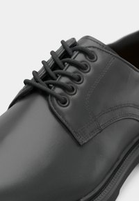 Chaussure habillée en cuir noir brillant, à laçage fermé et lacets ronds noirs passés à travers cinq œillets de chaque côté.