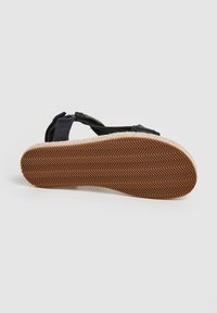Musta espadrille-sandaali, jossa on letitetty juuttipohja, varustettuna kumipohjalla, jossa on herringbone-kuvio, säädettävä hihna ja teksturoitu päällinen.
