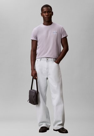 Ung mann iført en lys lilla Calvin Klein t-skjorte, hvite vide jeans, svarte sko, holder en liten svart skulderveske.