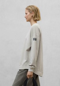 Sweatshirt gris clair avec une coupe décontractée, col rond et poignets côtelés. Comprend une étiquette noire sur la manche gauche et un tissu lisse et texturé.