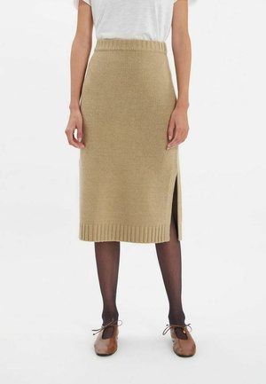 Jupe mi-longue en tricot beige avec taille côtelée et fente latérale, associée à des chaussures marron et des collants foncés. Design simple et texturé.