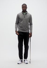 Άνδρας που στέκεται με μπαστούνι του γκολφ, φορώντας γκρι πουκάμισο με μακριά μανίκια, μαύρο παντελόνι και λευκά παπούτσια γκολφ σε απλό λευκό φόντο.