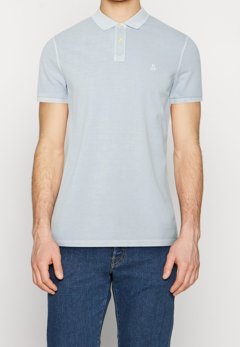 Homme portant un polo à manches courtes bleu clair à deux boutons et un jean bleu foncé, debout devant un fond uni.