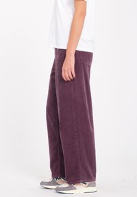 Pantalon large en velours côtelé d'un violet profond, avec une texture côtelée verticale, des poches latérales et une coupe décontractée, associé à des baskets neutres.