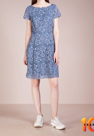 Kvinde iført en knælang blå blomstret blondekjole med korte ærmer og hvide chunky sneakers, stående mod en ensfarvet beige baggrund.