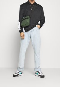 Chemise noire à manches longues avec accents blancs, jean baggy bleu clair, sac bandoulière vert, et baskets noires avec détails turquoise.